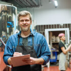 Ein lächelnder Brauer mit Klemmbrett und Schürze steht in einer Brauerei vor großen Edelstahltanks.