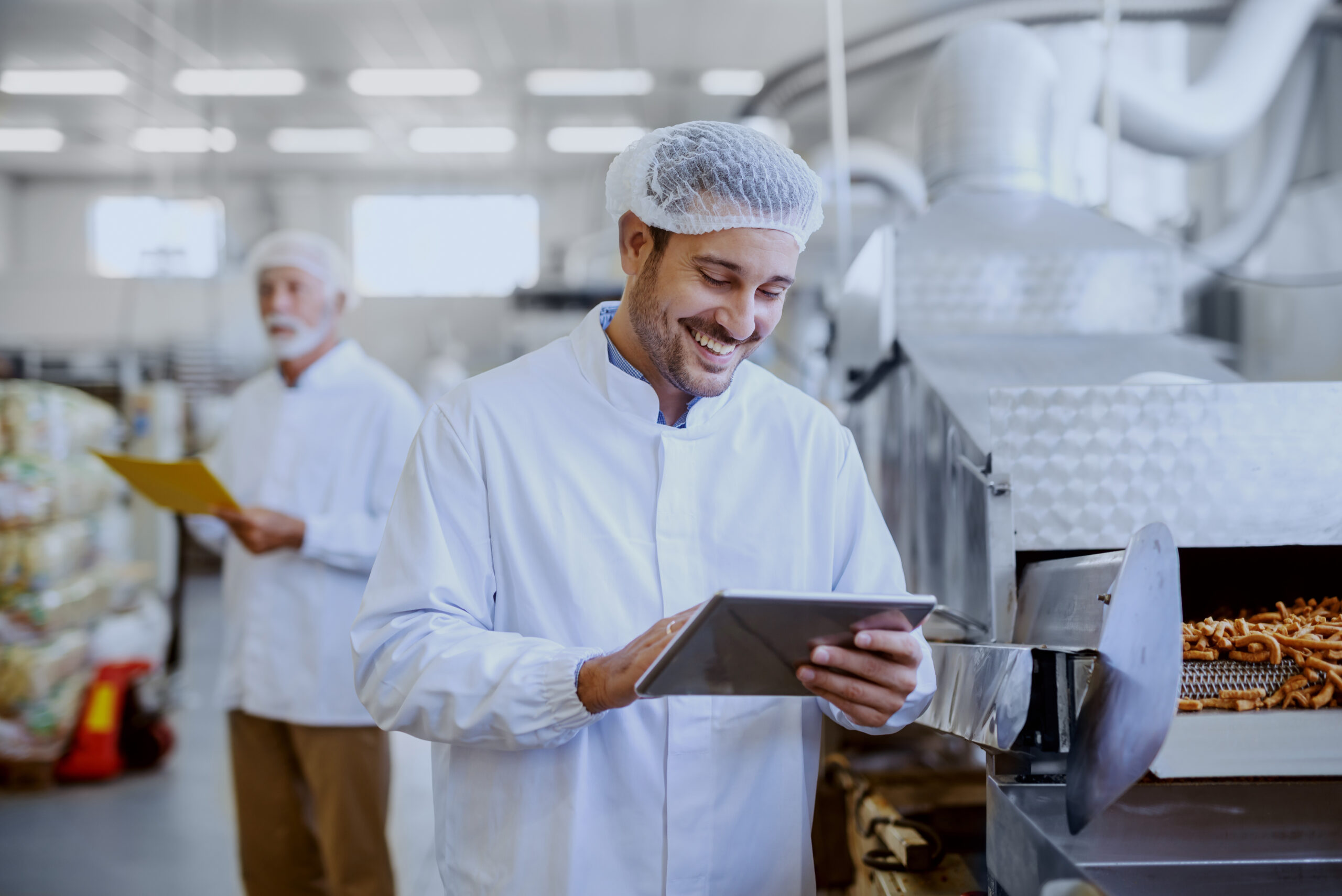 Young smiling supervisor in sterile white uniform using tablet and checking quality of salty sticks. In background older supervisor holding folder with documents. Food plant interior.